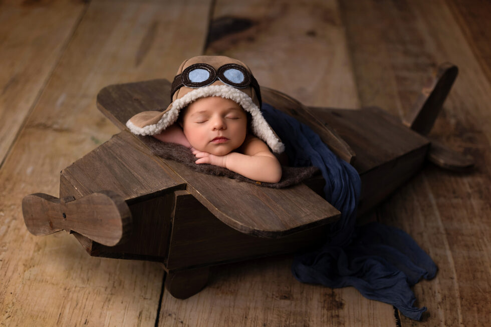 Bebé recién nacido dormido con gorro de aviador en un avión de madera, fotografía recién nacido artística en estudio en Getafe, Madrid