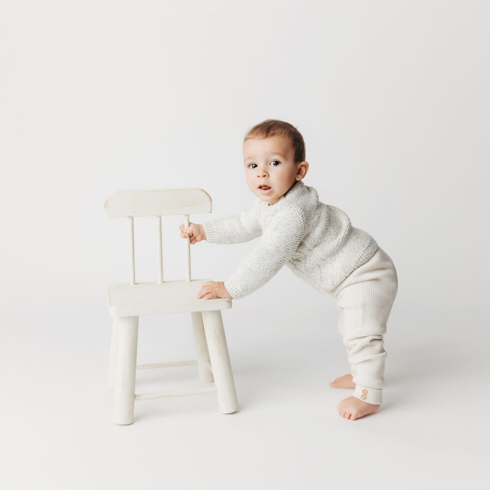 bebé de pie apoyado en una silla durante una sesión de fotografía infantil en estudio